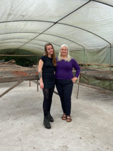 Two woman, one with dark hair and one with blonde hair, stand in a greenhouse in front of drying racks full of cacao beans.