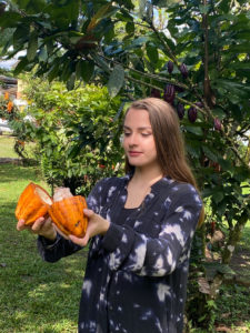 Young woman with dark hair holding an orange colored cacao bean pod that is split open.