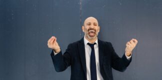 Bald man with dark and gray facial hair wearing a dark suit and tie with a white shirt. His mouth is wide open and he appears to be speaking while making hand gestures