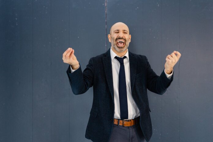 Bald man with dark and gray facial hair wearing a dark suit and tie with a white shirt. His mouth is wide open and he appears to be speaking while making hand gestures