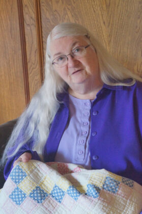 Women wearing blue and wire-rimmed glasses with long white hair. Her head is cocked like she's listening intently.