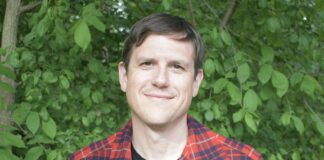 John Counts is an author based in Michigan. He is shown here from the waist up. He has short brown hair and is wearing a black t-shirt with a red and black flannel over the top. There is leafy foliage in the background.