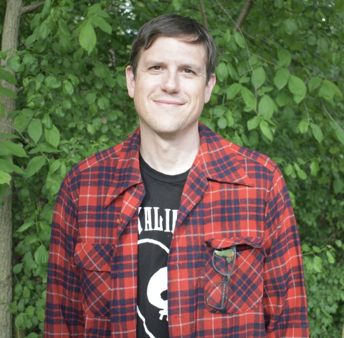 John Counts is an author based in Michigan. He is shown here from the waist up. He has short brown hair and is wearing a black t-shirt with a red and black flannel over the top. There is leafy foliage in the background.