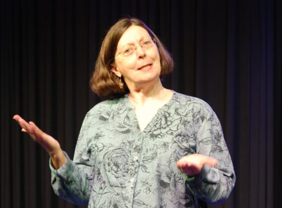 Woman with dark hair in a bob wearing wire-rimmed glasses and a gray shirt with black pattern uses expressive hand gesture and appears to be talking