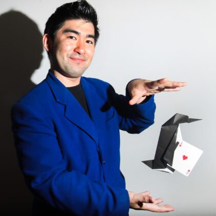 Man with short dark hair wearing a blue shirt seems to magically levitate an origami penguin