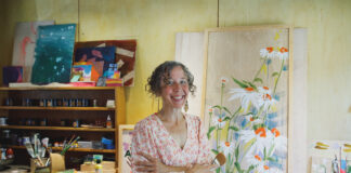 Popular Ann Arbor, Michigan artist, Anne Erlewine, has her arms crossed over her body and is wearing a colorful flowered shirt with a white background and orangey flowers with a light blue skirt. She has curly, graying hair that is pulled back. She is standing in her studio with art supplies and a large painting of daisies in white, blue, green, and orange propped up in the background