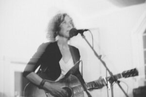 Black and white image of popular Ann Arbor, Michigan musician Anne Erlewine singing at the mic as she plays the guitar. She is has curly graying, shoulder-length hair and is wearing a black blazer with a white shirt underneath.