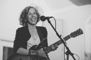 Black and white image of popular Ann Arbor, Michigan musician Anne Erlewine smiling at the mic as she plays the guitar. She is has curly graying, shoulder-length hair and is wearing a black blazer with a white shirt underneath.