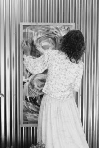 Black and white image of the back of popular Ann Arbor, Michigan artist Anne Erlewine hanging and abstract painting, that is mostly blocked by her body, on a striped wall. She has curly, shoulder-length, graying hair and is wearing a white patterned long-sleeve shirt with a light-colored skirt.