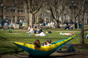 Two students in a yellow and blue hammock look out over the lawn near The Diag, which is filled with small groups of students laying out, sitting in the grass, and enjoying the sun.